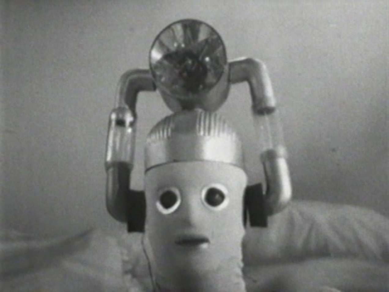 Image: A Cyberman looks directly into the camera. It has a large lamp attached to its head via tubes connected to the ears. There is fabric stretched over its face, with rounded eye-holes and a slit for a mouth.