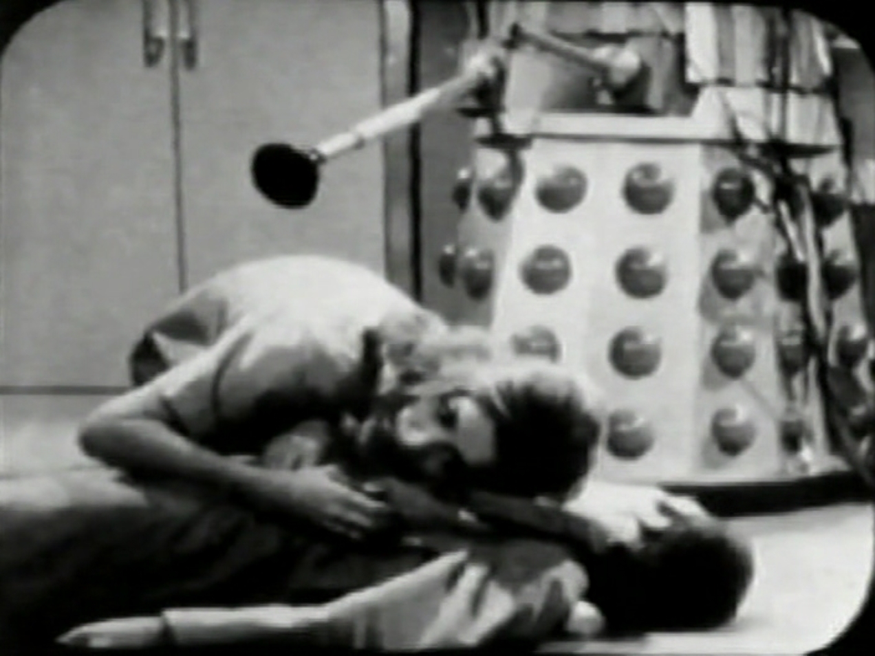 A man lies unconscious on the floor. A woman, Janley, listens to his chest. There is a Dalek looming over them. 