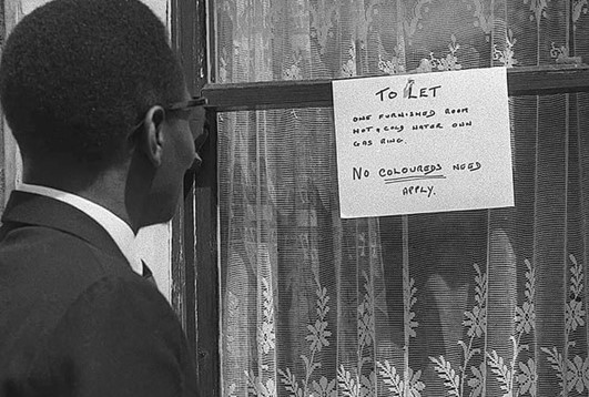 Black Man looking at sign in window saying: "To Let: No Coloureds Need Apply" 