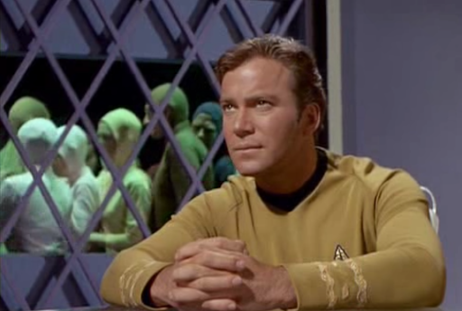 Kirk looks sternly at Ambassador Hodin offscreen. In the background, we see the people of Gideon milling around aimlessly.