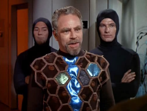 Ambassador Hodin wearing a suit mostly made of brown velvet hexagons with some kind of wide ribbon between them, and a shiny metallic blue row down the front. He is flanked by two assistants in all-black hooded bodysuits.