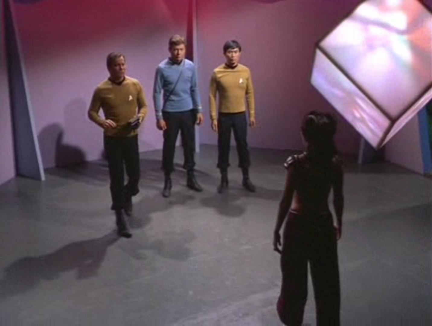 Elevated shot of Kirk, McCoy, and Sulu in an octagonal room, Losira in front of them, a shimmering cube on the ceiling in the upper right