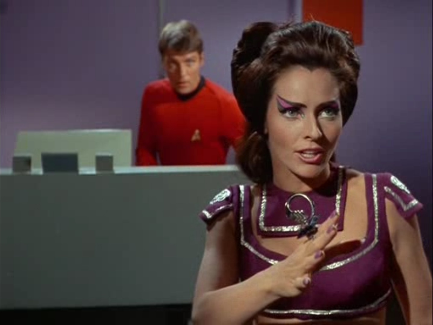 Losira, in a purple outfit, stretches her hand out toward the party as it begins to beam down, a transporter technician in the background