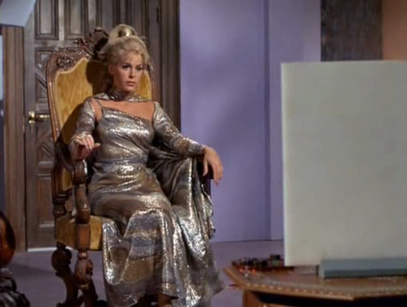 Rayna, sitting in a chair and wearing a polychrome, metallic gown, views a cream-colored flat screen