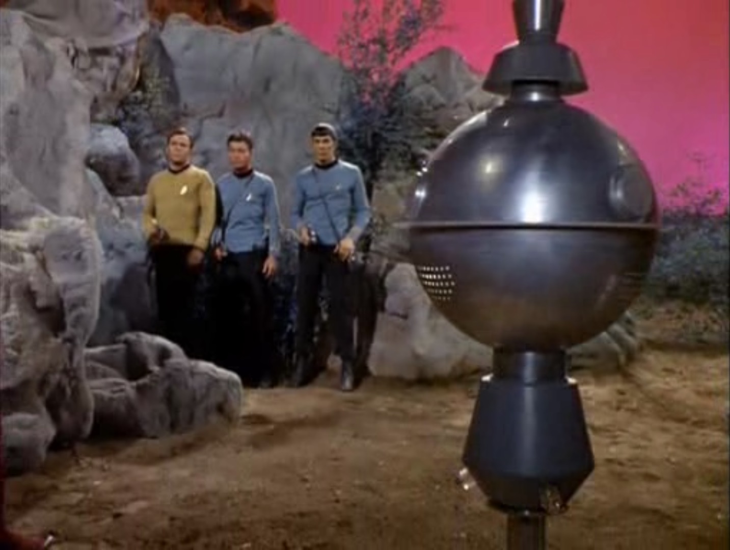 A spherical robot of gray steel floats menacingly in front of Kirk, Spock, and McCoy, who have their phasers out