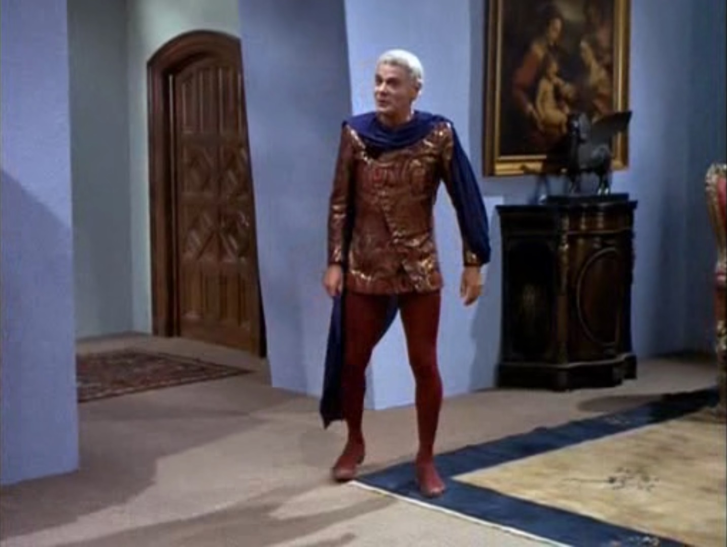 Image of Flint, an older man in a ceasar hair cut, a futuristic Shakespearian noble outfit, complete with tights, standing in a blue-walled room with a Renaissance painting on the wall