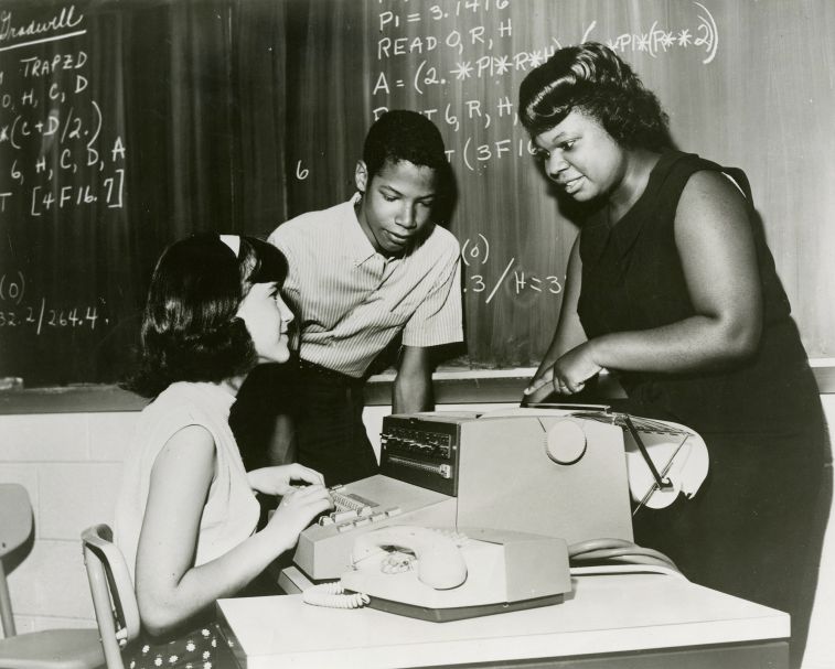 black and white photo of a plump Black woman leaning over an eighth-grade white girl seated at a computer, a eight-grade black boy behind her, mathematical equations on the blackboard behind them all