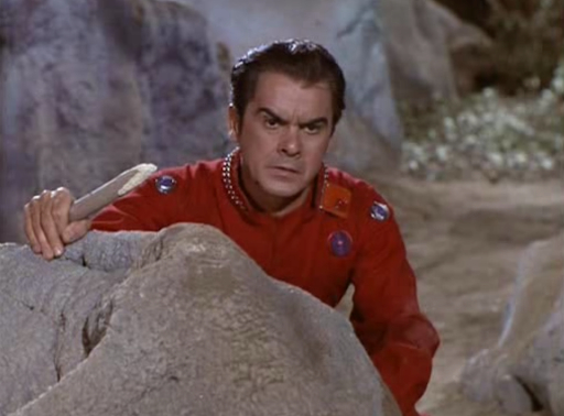 screen cap of Colonel Green, a swarthy middle-aged man in a red jump suit holding a sharpened stick taking cover behind a styrofoam boulder