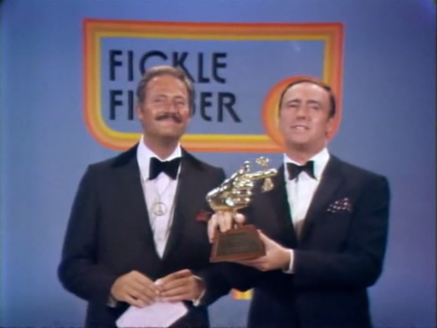 photo of a two men in suits (Dan Rowan and Dick Martin), the one on the right holding up a golden statuette of a hand with its index finger pointing and crowned by wings