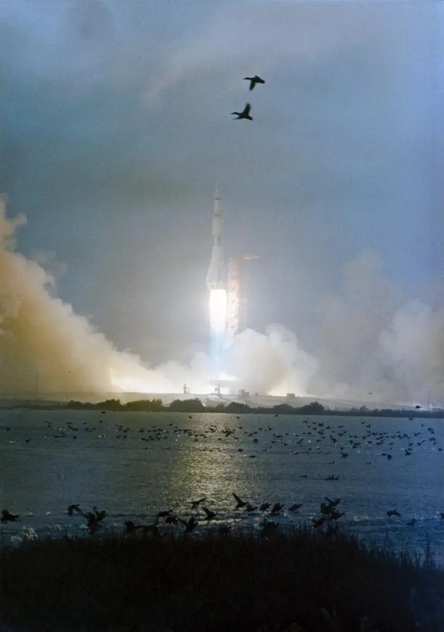 A color photograph of Apollo 12's Saturn V lift-off from Kennedy Space Center. The sky is completely overcast. The fuel burning at the back of the launcher makes a bright spot in the center of the photograph, with fumes and steam on the sides of the launchpad. Two birds are passing in the frame, near the photographer.