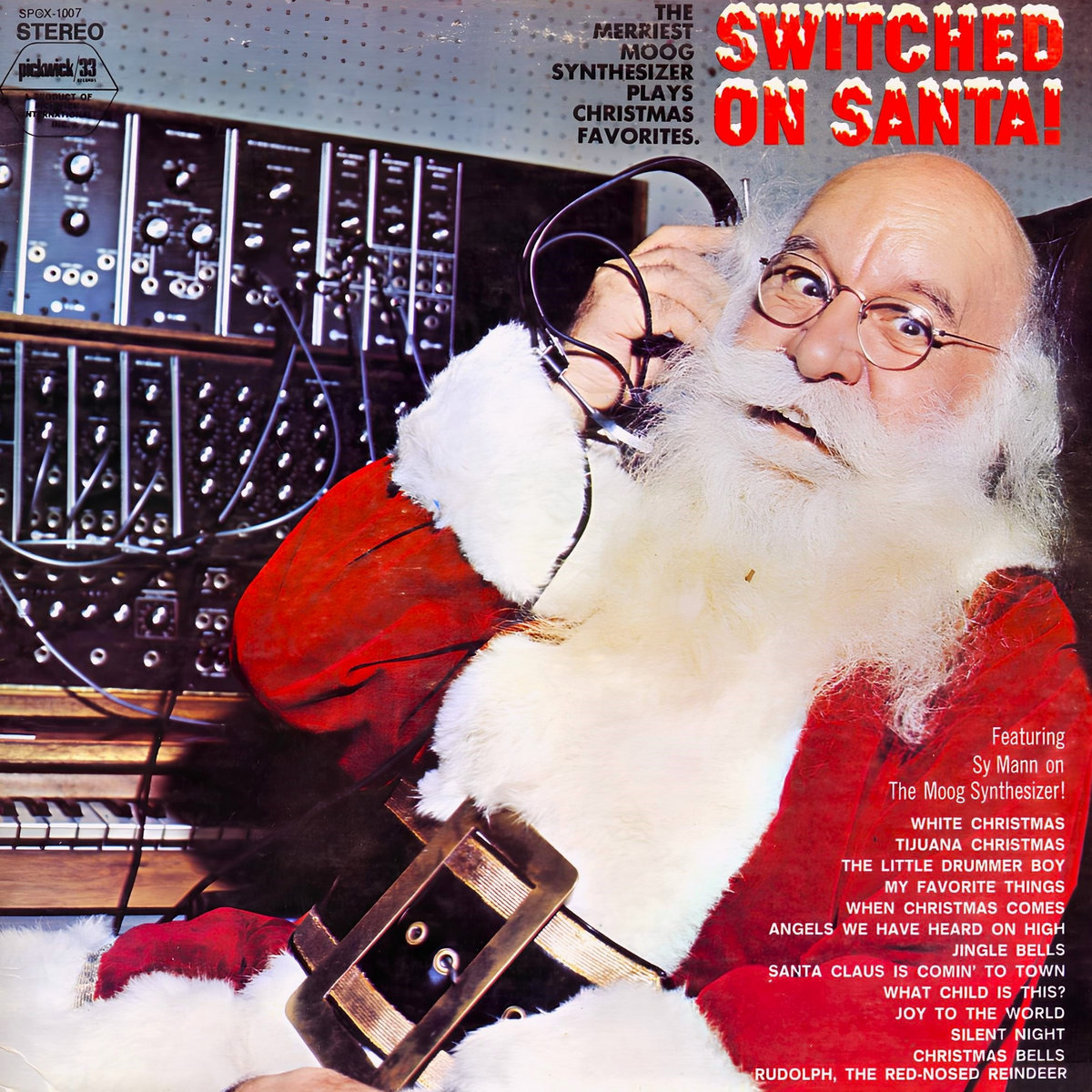 Vinyl record jacket, titled Switched on Santa! The merriest Moog synthesizer plays Christmas favorites. Text at the bottom right corner says: Featuring Sy Mann on the Moog synthesizer! Under this line, the text announces the songs White Christmas, Tijuana Christmas, The Little Drummer Boy, My Favorite Things, When Christmas Comes, Angels We Have Heard On High, Jingle Bells, Santa Claus Is Comin' to Town, What Child Is This, Joy to the World, Silent Night, Christmas Bells, and Rudolph the Red-Nosed Reindeer. The image on the record jacket is a photograph of Santa Claus sitting next to specialized equipment in a sound recording studio, holding an end of a headphone diadem next to his right ear.