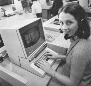 photo of a young brunette woman, sitting at a computer and wearing a headset. She is wearing a short-sleeved ribbed sweater, and is smiling over her shoulder at the camera.