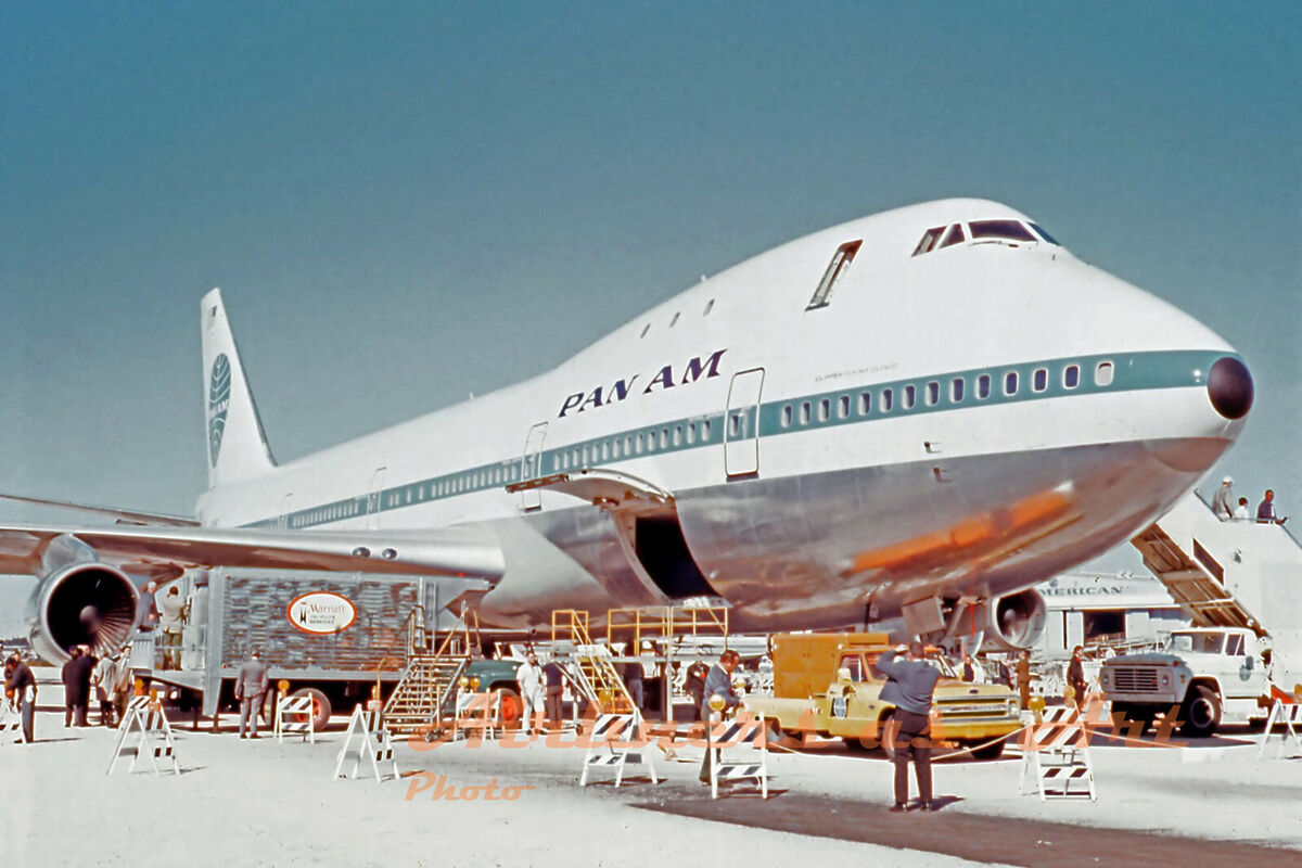 photo of an enormous jet, parked on the ground on a sunny day. There are also observing members of the public, of which there seem to be about 4. The top half of the jet is white with a horizontal turquoise stripe that extends all the way around. Above the stripe, there are the words PAN AM in large black letters. The bottom half of the plane is polished, reflective metal, and there is an open hatch on the left side, closest to the photographer. On the right side of the image, we can see the stairway allowing passengers to depart. On the left side of the image, there is a small barrier of folding wood signs between the photographer and the jet. The barrier surrounds a group of 3 trucks and 8 or so technicians, as well as the platform ladders that reach from the ground to the open hatch.