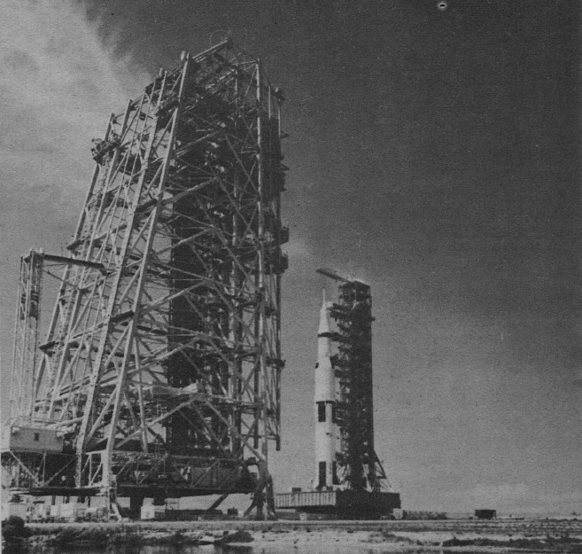 photo of a gantry tower constructed of metal struts. In the background, there is the Saturn V, attached to a similar structure. They are both on large platforms that seem to float above the ground.