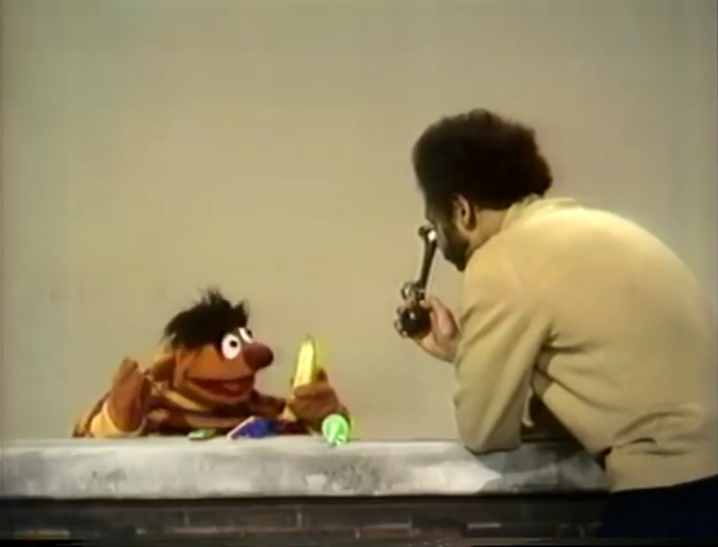 still photo of a Black man (Gordon) proffering a bicycle horn to an orange puppet with unkempt hair (Ernie) holding a banana