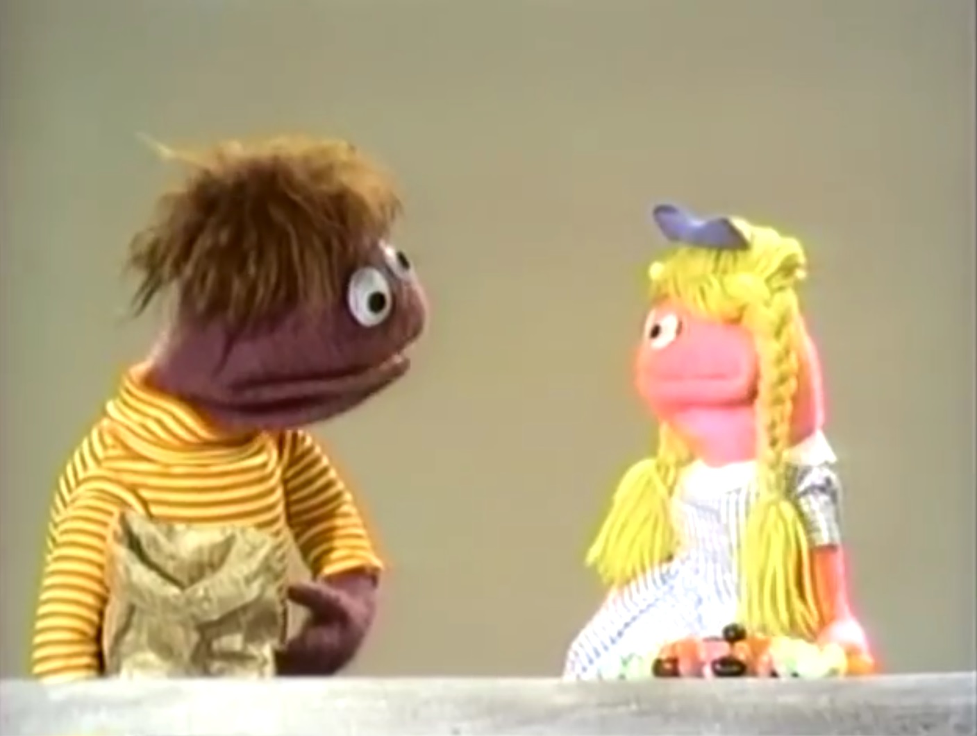 still photo of two puppets facing one another, one with brown paper sack, and the other with a pile of jelly beans