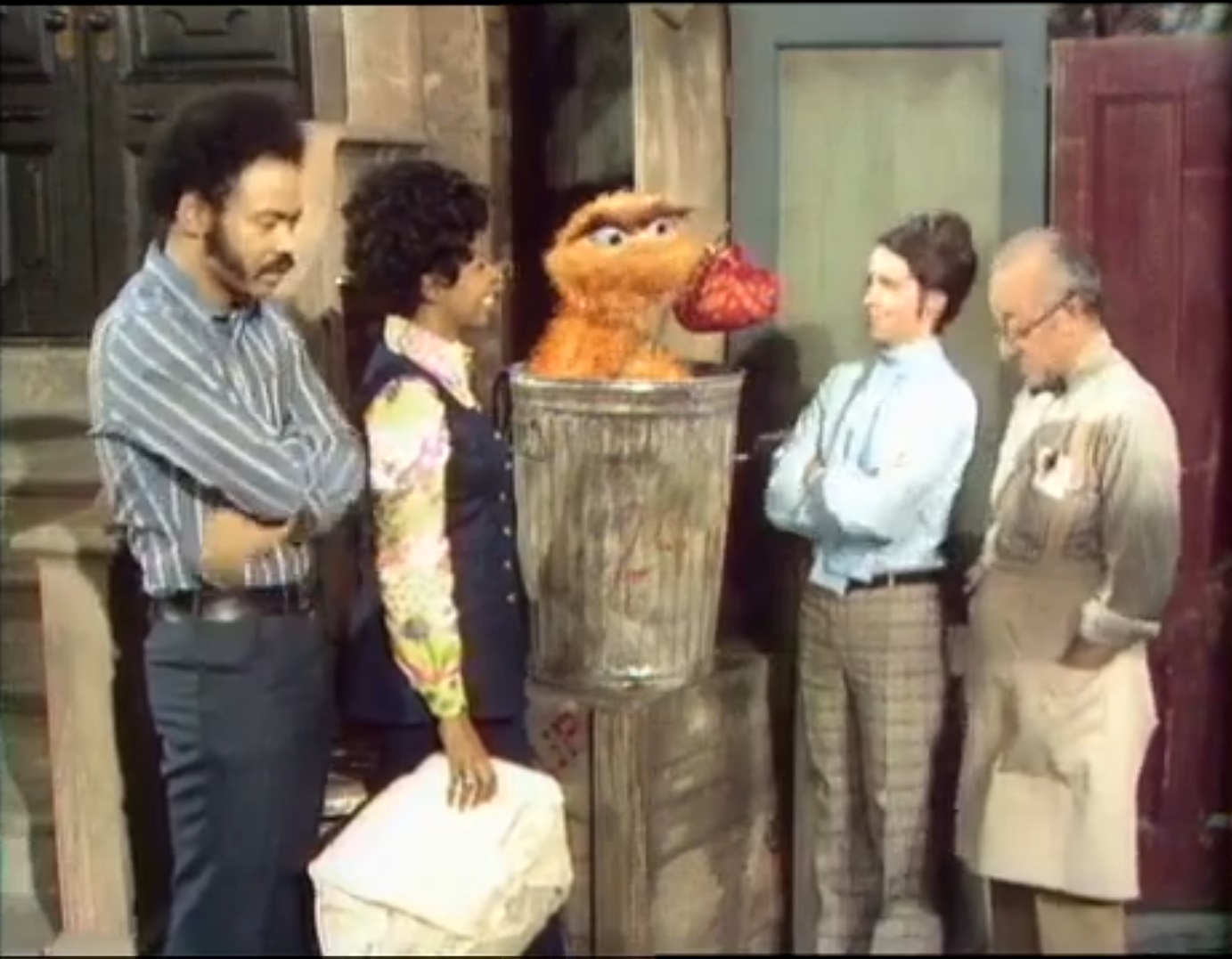 still photo of a graying White man in a shopkeeper's apron (Mr. Hooper), a White woman (Jenny), a Black woman (Susan), and a Black man (Gordon) all flanking an furry orange puppet with a hobo's bindle