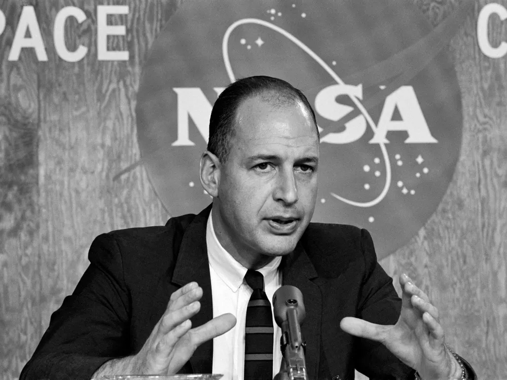 Black and white photo of Apollo manager George Low speaking into a microphone in front of a NASA press backdrop.