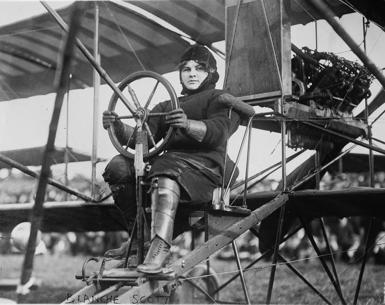 Photo of a cold-weather suited young woman behind the wheel of a Curtiss Model D, open-air biplane