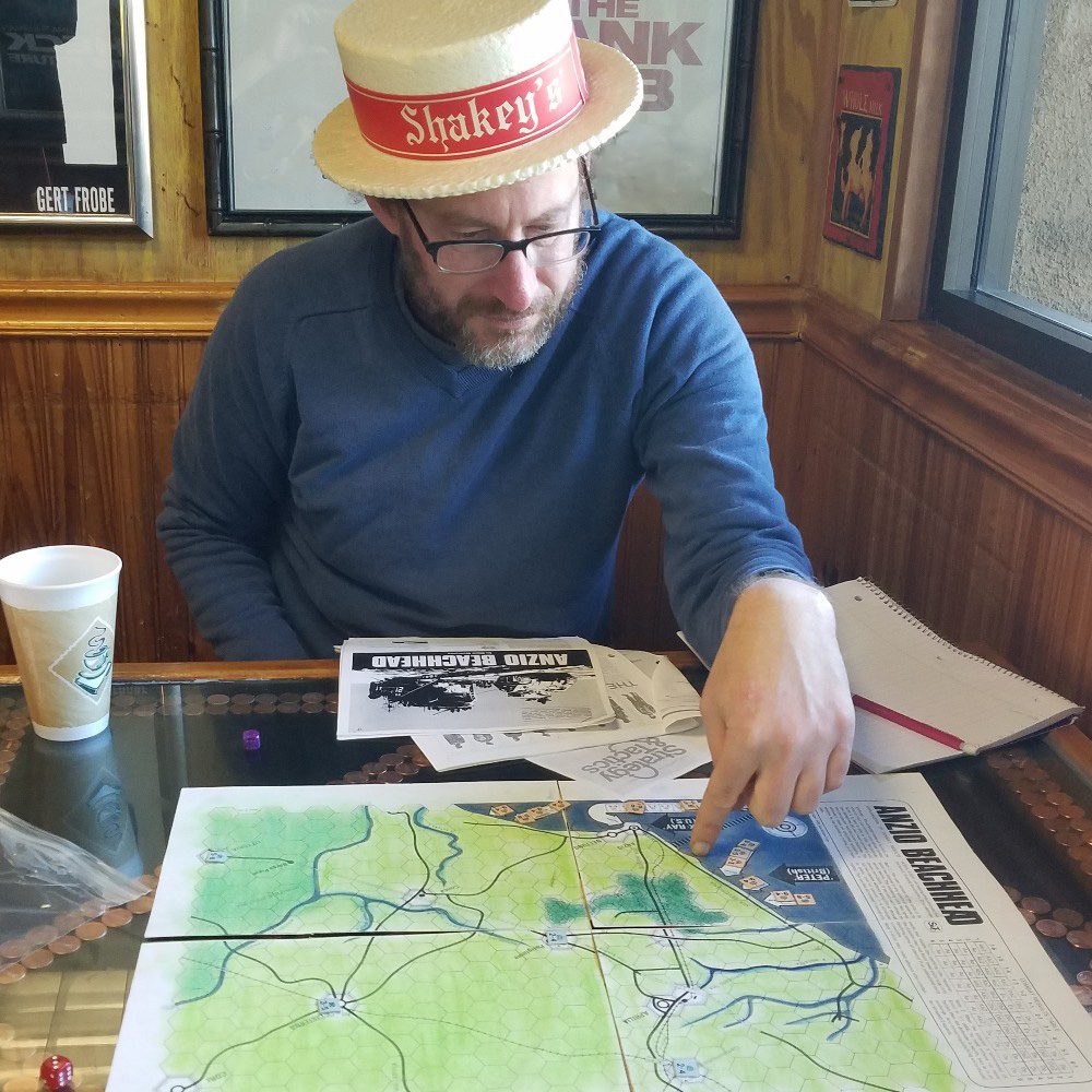 Gideon, wearing a Shakey's faux straw hat, points at the Anzio Beachhead map in this photo, taken in a diner.