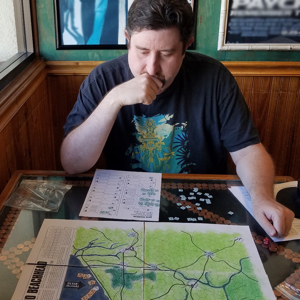 Perfect John, a white man with a pensive expression, contemplates the Anzio Beachhead map in this photo. He is seated in the corner of a diner.