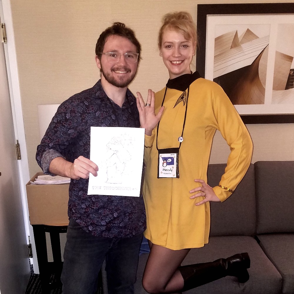 Photo of a bearded man in glasses and a paisley shirt holding up a copy of a fanzine next to a tall woman in a Trek gold tunic flashing the Vulcan salute