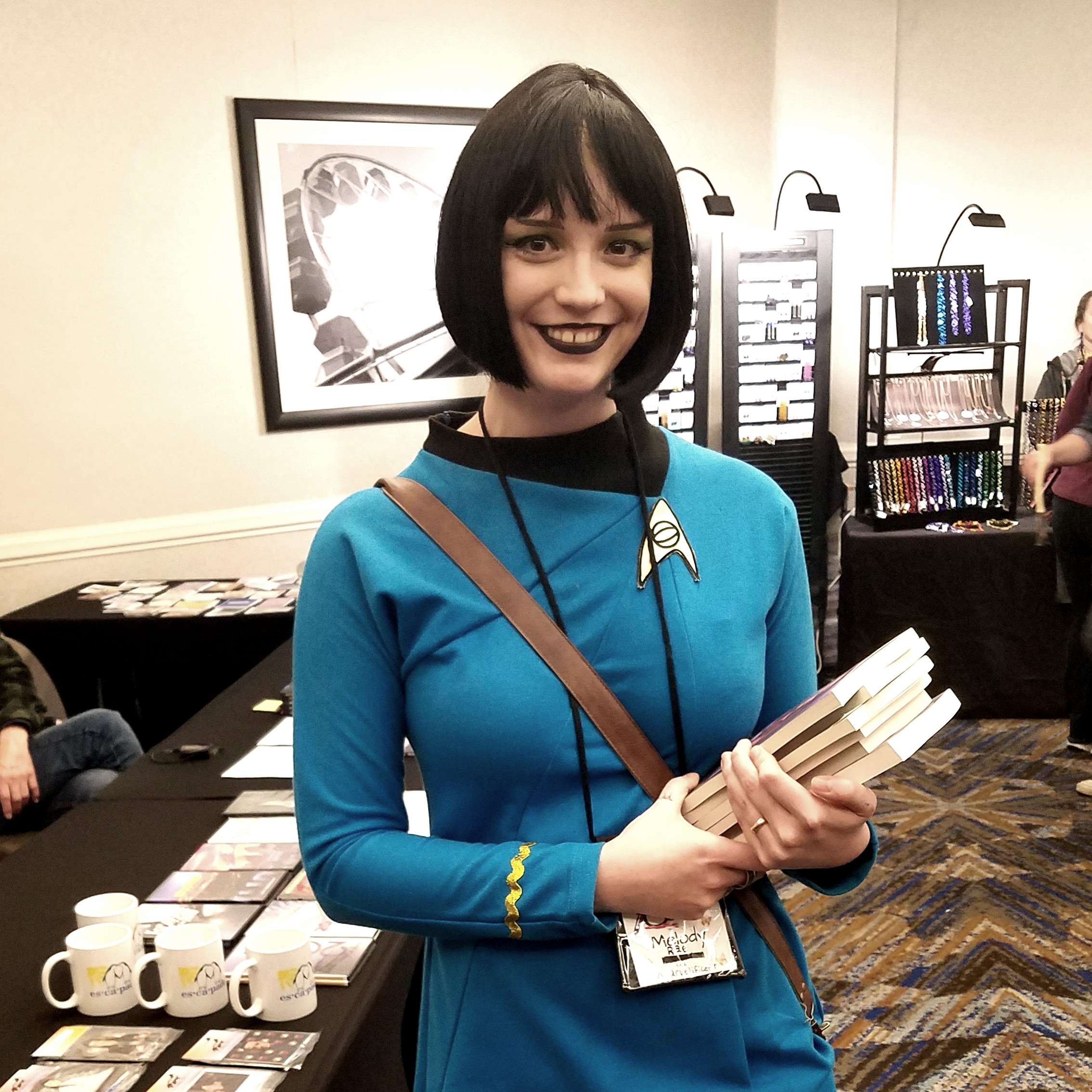 Photo of a dark-haired woman in a blue Star Trek uniform, smiling at the camera. She is carrying books in one arm, and behind her are tables of fannish items for sale.