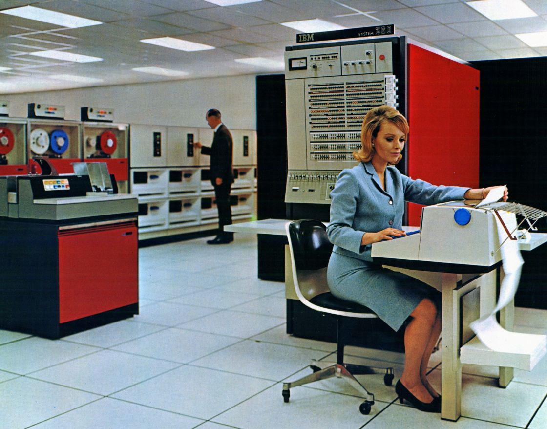 A woman in a blue dress, sitting at a printer desk. In the background, a man in a dark suit is punching into a large computer.