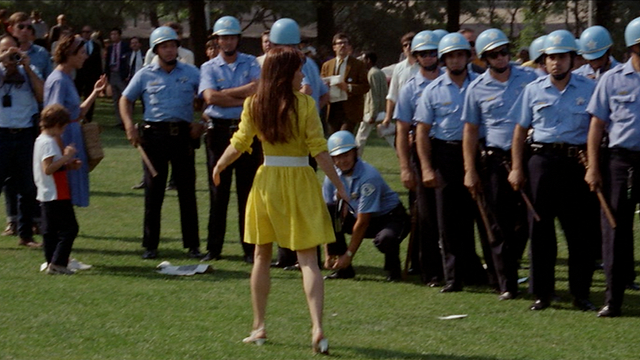 A brunette woman in a yellow dress walks in front of a line of police officers. There are hippies of various ages at the left side of the frame, including a young boy.