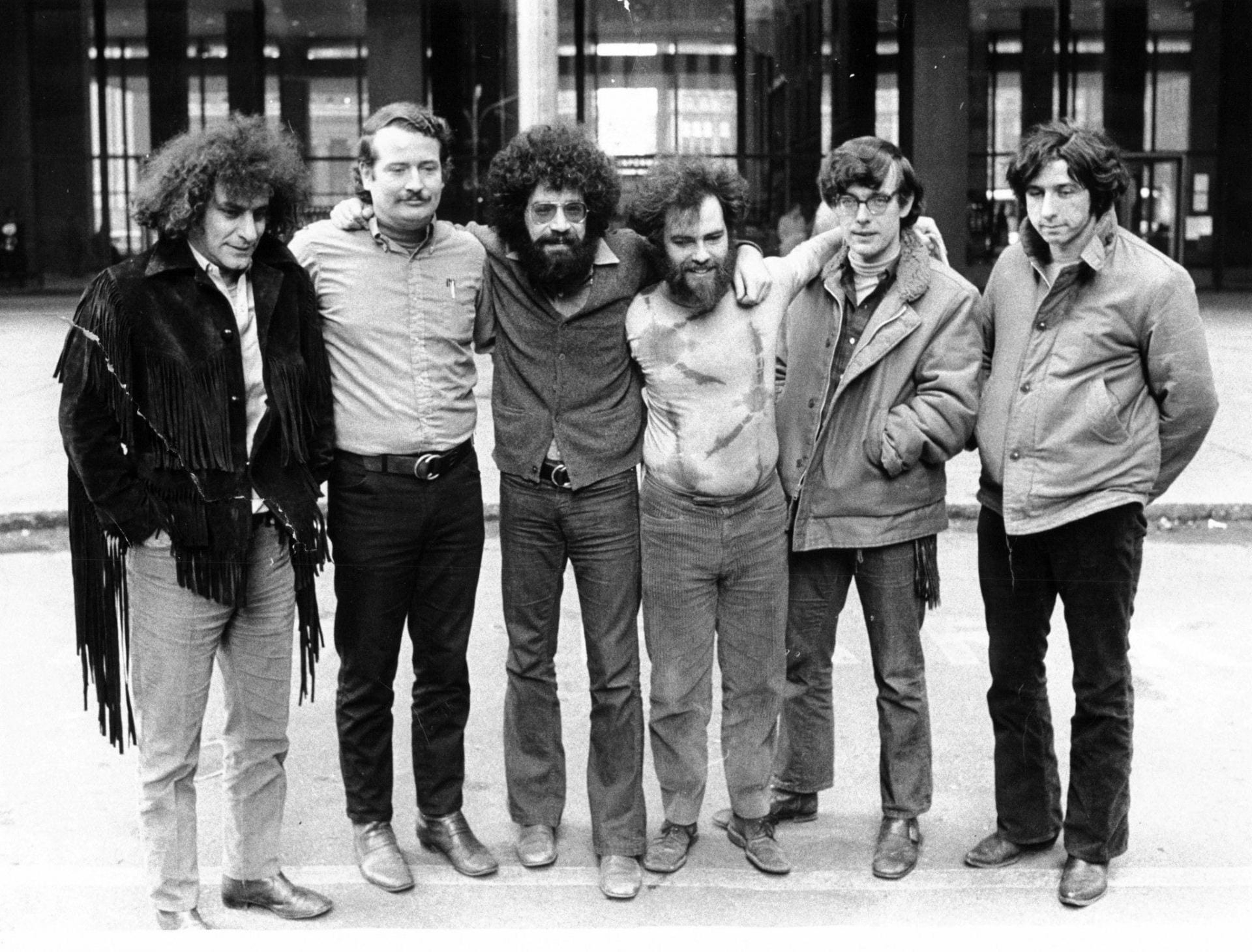 Six men with long, wild hair stand smiling at the camera, arms wrapped around each other