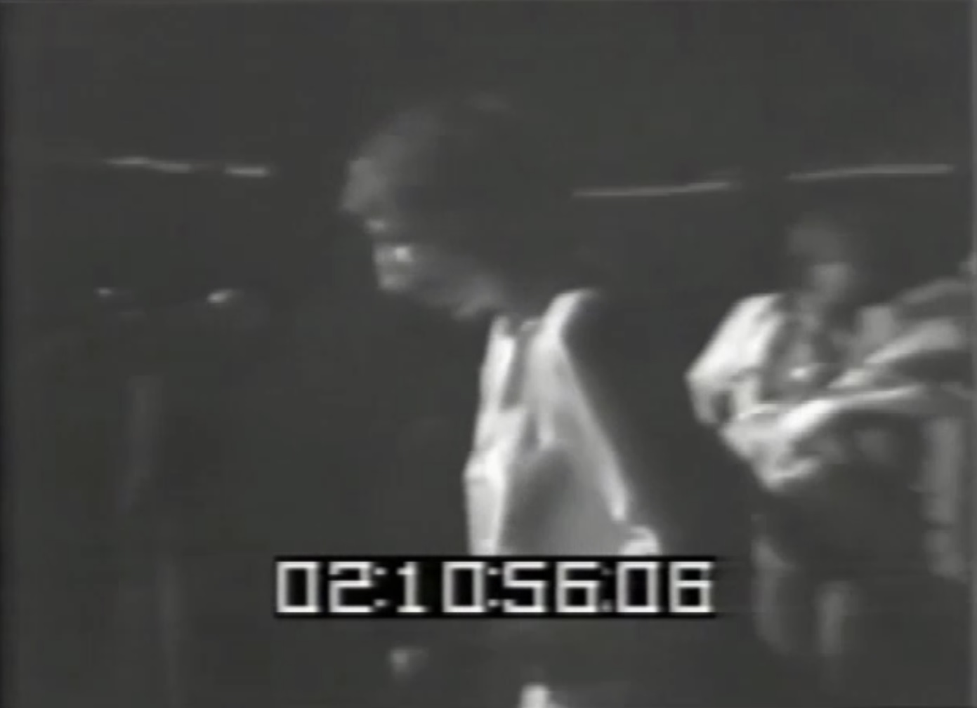 A blurry black-and-white still of a floppy-haired white man in a white tunic. The image is timestamped 02:10:56:06