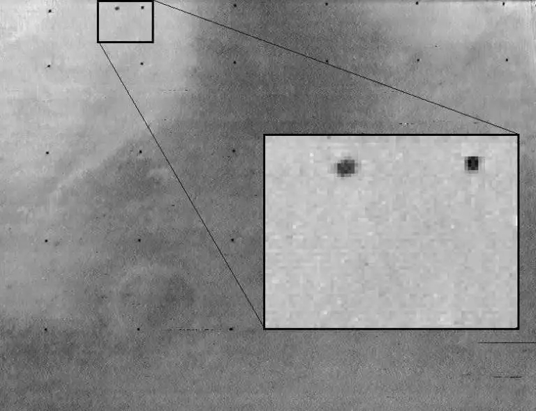 Very blurry black-and-white photograph of the Martian moon Phobos, visible only as a tiny smudge of irregular shape against a gray background.