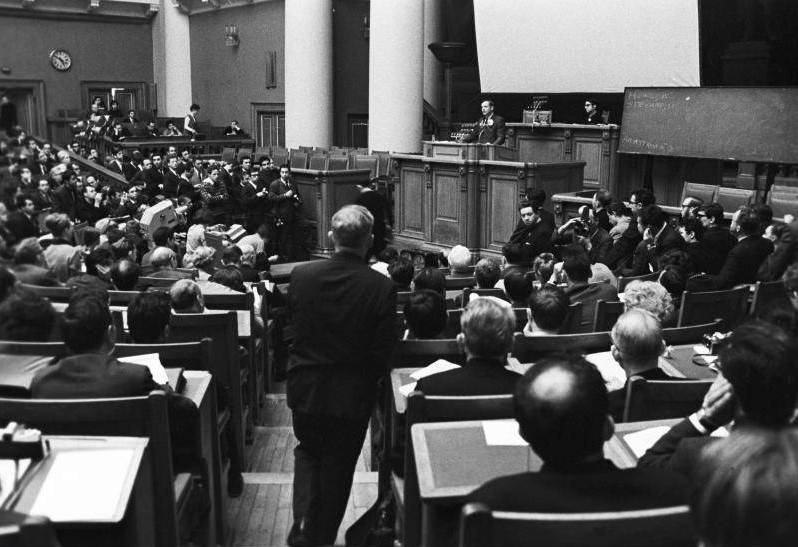 A black and white photo of a full lecture hall.  The chairs and desks are arranged in a half circle around a central podium, where two men sit facing the crowd.