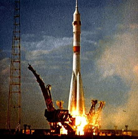A color photograph shows a white cylindrical rocket with a thick red stripe around the middle against a cloudy blue sky.  Four metal support structures are falling away from the rocket as the thrusters ignite. On the left of the frame a signal tower extends upward out of the frame. 