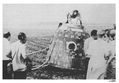 A black and white photograph shows the dome-shaped crew compartment of the Soyuz after landing in a field. It has several metal legs sticking out at odd angles from teh sides and bottom.  Several men are gathered around the compartment, watching as an astronaut climbs out of the top.