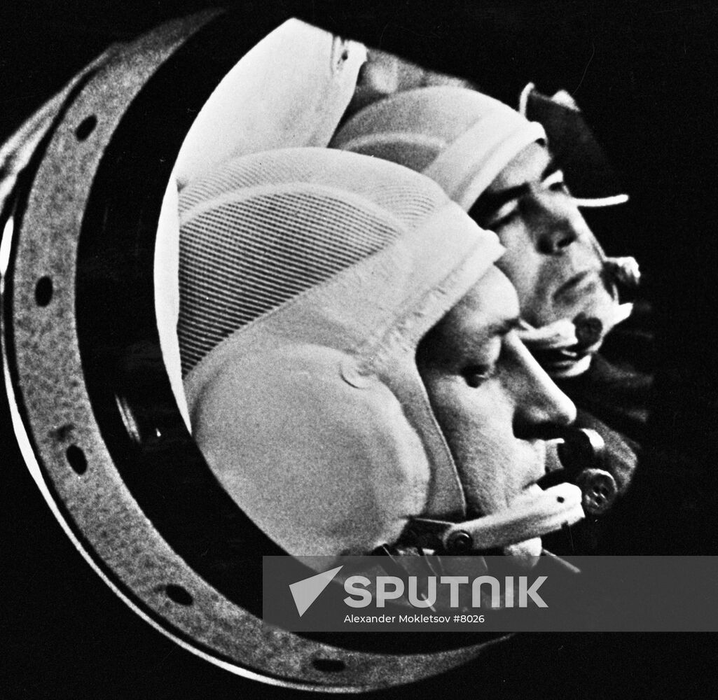 A black and white photograph of a round viewport, through which the heads of two white men are visible.  They are wearing headgear with a mesh scalp, thick ear coverings, and a headset microphone extending in front of their mouths.  The caption at the bottom right says Sputnik.