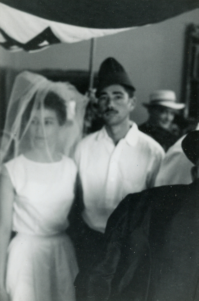 Very blurry black-and-white photograph of a couple at their wedding. The bride is wearing a translucent veil and a slim, sleeveless, unadorned white dress. The groom is wearing a plain white shirt, black pants, and a conic hat.