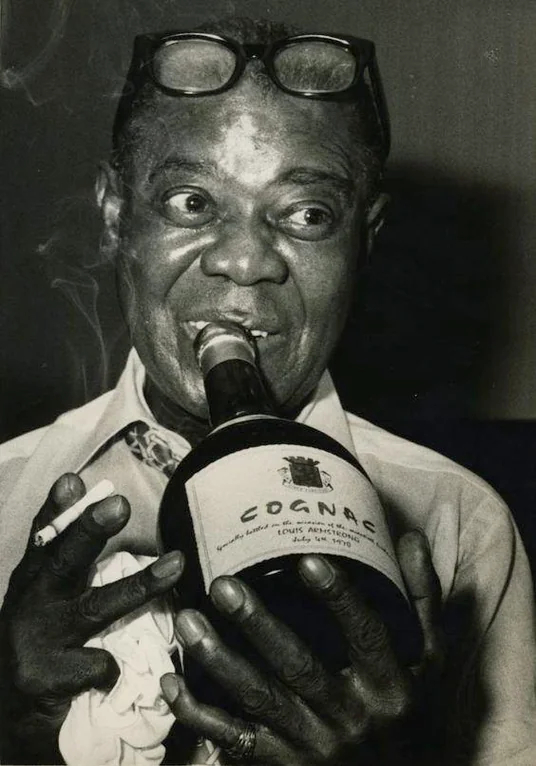 Photo of a smiling Louis Armstrong, with a cigarette and a bottle of cognac.