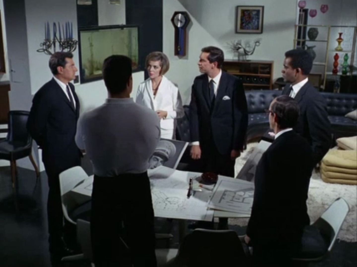 A color photo of several men and a woman, all in business attire, standing around a conference table in an office.