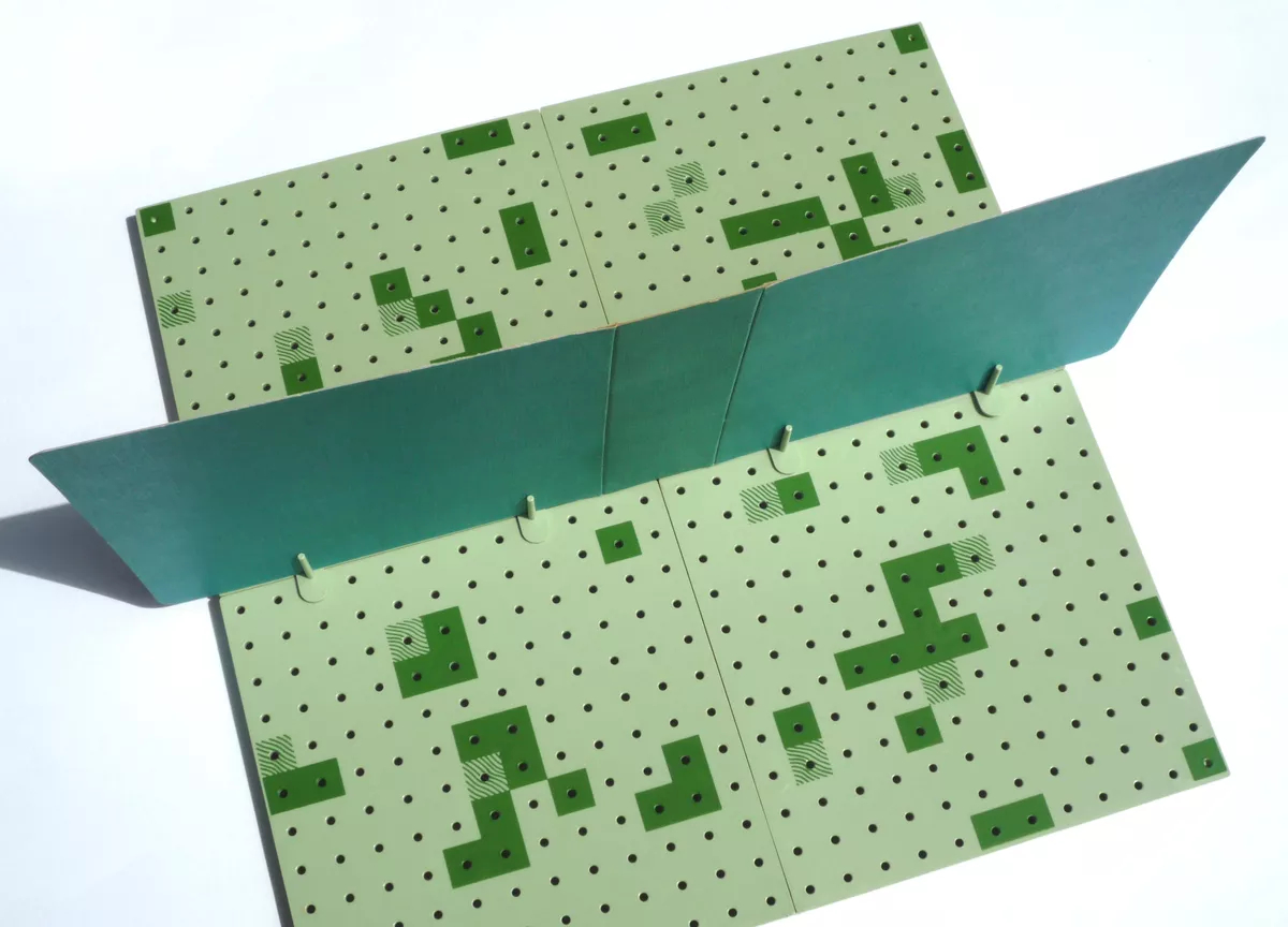 Colour photograph of the light-green game board from above.  Scattered across the squares of the board are variously configured groups of contiguous squares in either solid (dark) green or with green wavy lines.  A vertically screening divider is positioned across the center of the board