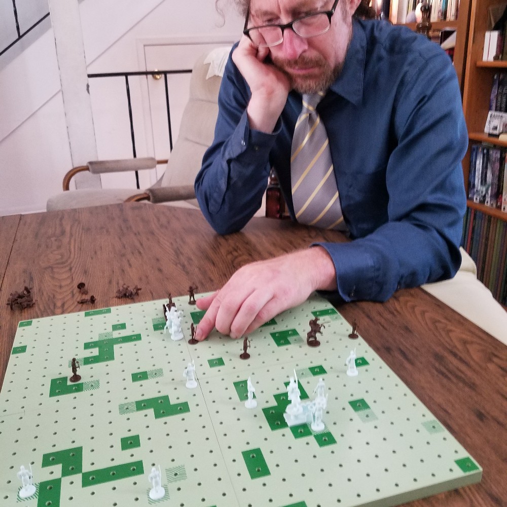 Colour Photograph of a seated white man with concerned expression reaching to move one of his remaining pieces