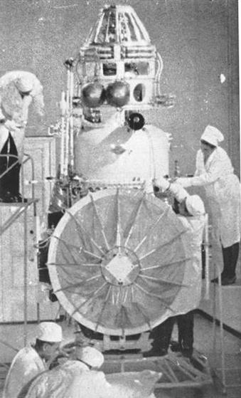 Black & white photograph of five white-coated engineers gathered around a all sides of a complex machine approximately four meters in height.  Various hoses and pressure chambers are in evidence around the probe's chassis, and there appears to be an umbrella-like dish facing towards the camera