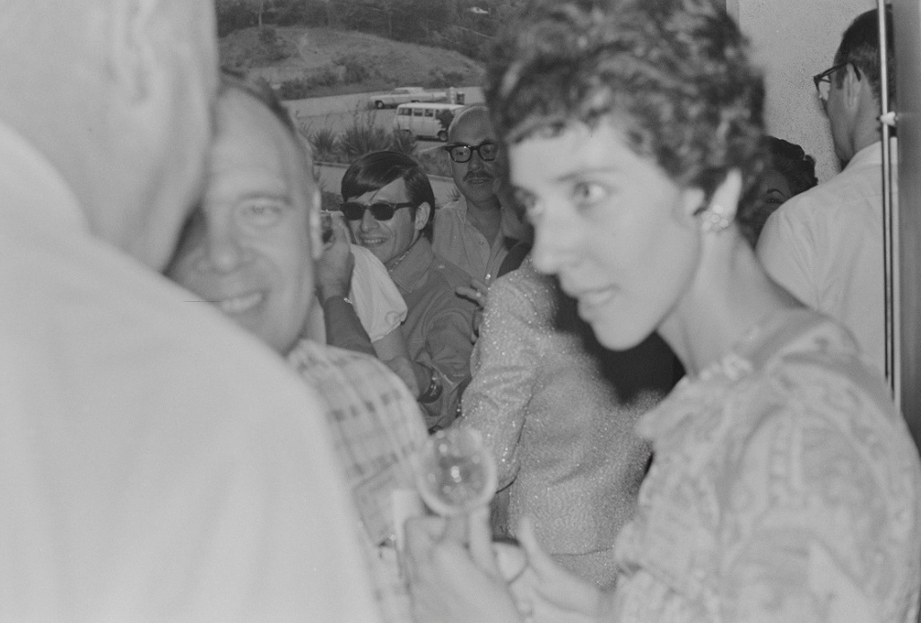 Black-and-white photograph of a woman with short hair talking to two men at a social gathering.