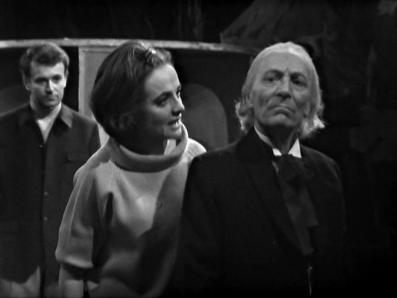 Image description: In the foreground, Barbara talks to the Doctor. Both appear angry. Ian glares at the Doctor from the background.