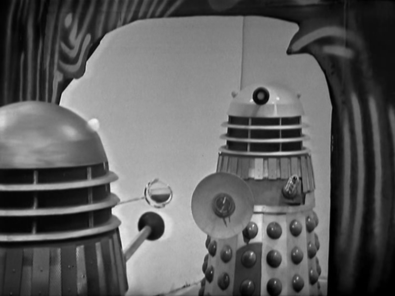 Image description: One Dalek in the foreground facing away from the camera. Another Dalek in the background facing towards the camera, standing in a lit doorway.