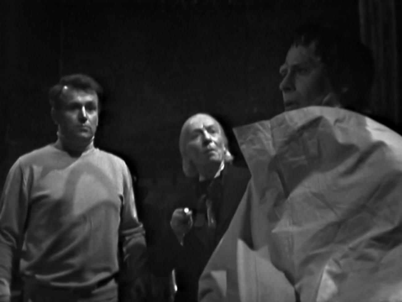 Image description: In the foreground, Frankenstein's Monster is sitting up, partially covered by the sheet. In the background, Ian and the Doctor regard him with apprehension.