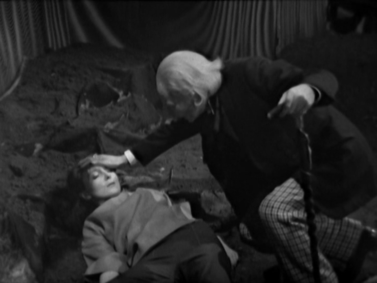 Image description: Vicki lies on the floor while the doctor crouches over her, feeling her forehead.