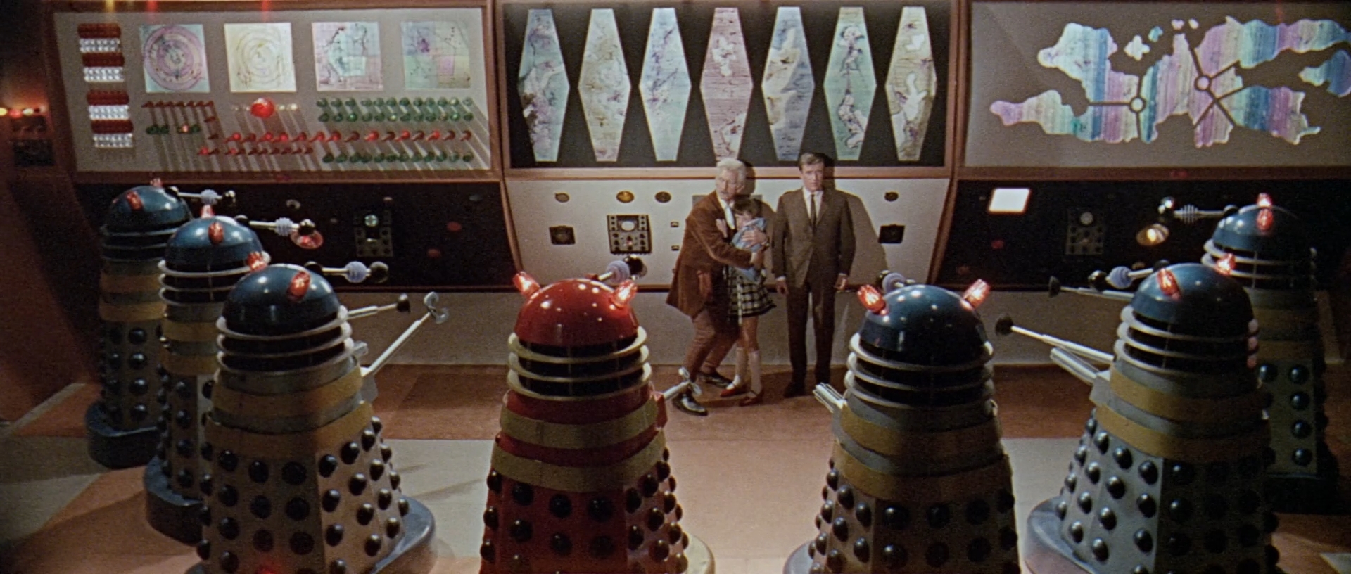 Image description: 7 Daleks in the foreground, looking at Dr. Who, Susan and Ian in centre frame. There is a computer bank in the background.