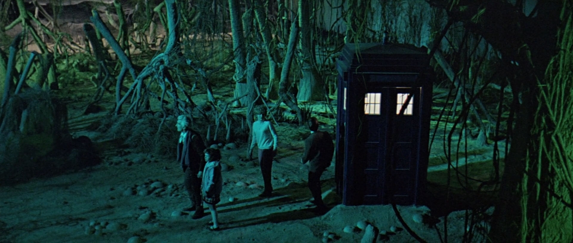 Image description: Wide shot of petrified forest in green lighting. The four main cast stand in centre frame, beside Tardis.