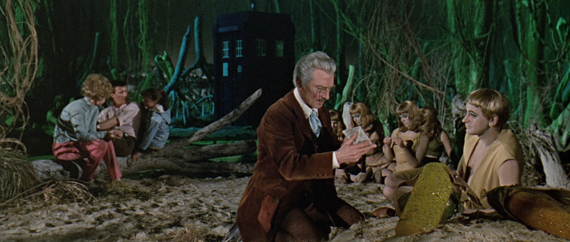 Image description: In the foreground, Dr. Who kneels with Alydon and examines some writing on a stone. In the background Barbara, Ian and Susan sit together. Tardis is visible in the distance.